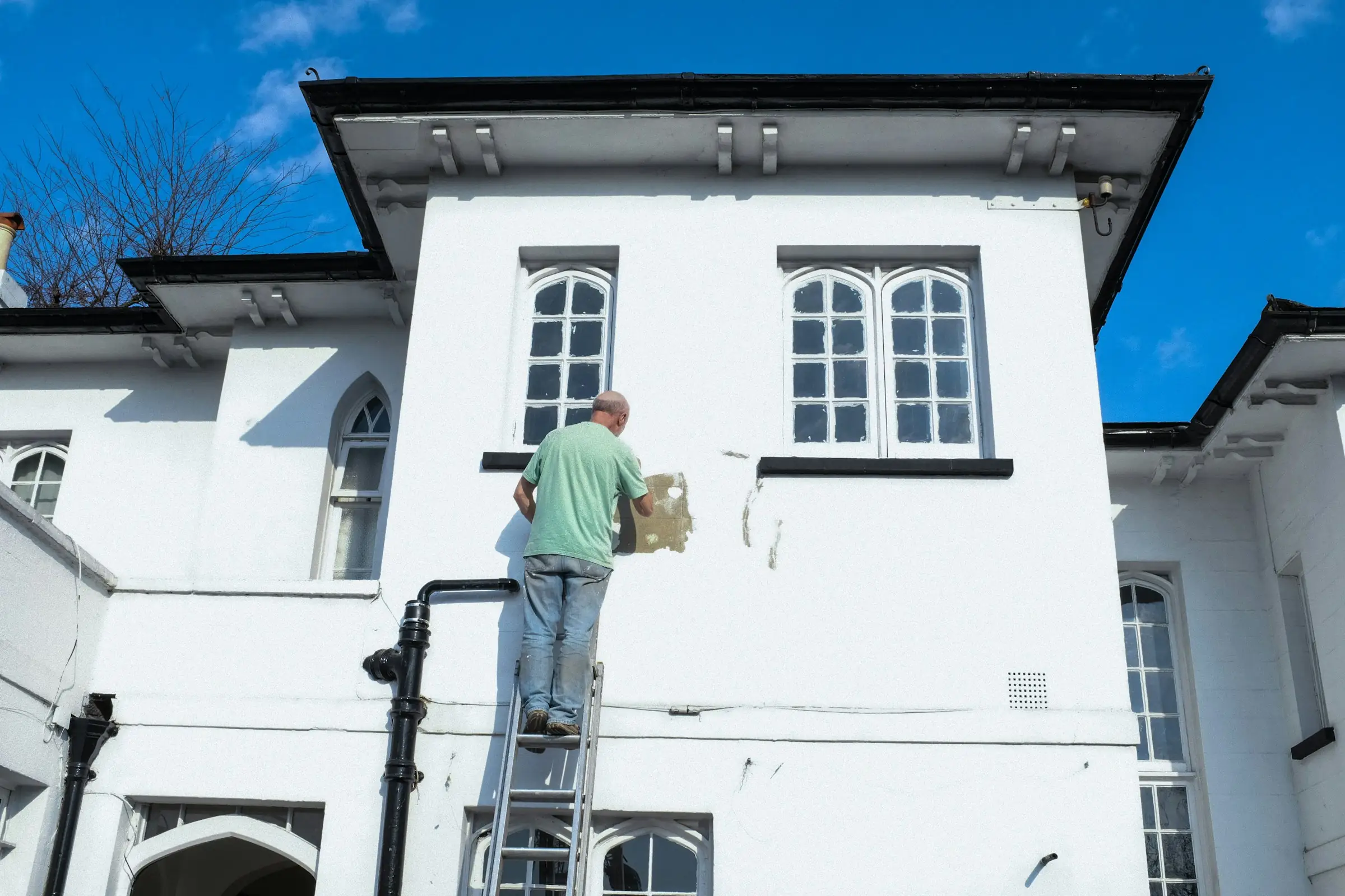Painter on a ladder preparing the exterior wall of a white two-story home